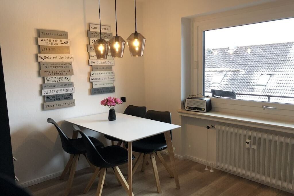 A dining area with a white table, black chairs, a vase with pink flowers, and a wall with various kitchen and family rules.