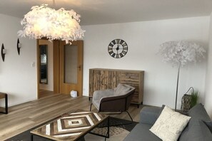 A modern living room with a wooden floor, a sofa, a chair, a patterned coffee table, a wall clock, and a decorative light fixture. - Holiday apartment with 1 bedroom (Hahnenklee Bockswiese)