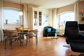 A room with a dining table, chairs, a black sofa, a cabinet with glass doors, and a window with curtains. - Residence Juliana 47 (Julianadorp)