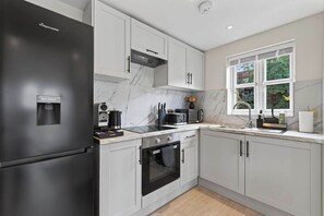 A modern kitchen with a black refrigerator, white cabinets, a built-in oven, and a window with a view of greenery. - New! Stylish 1BR Flat in Lewisham (Greater London)