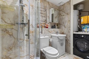A compact bathroom with a glass-enclosed shower, a toilet, a sink, and a washing machine. - New! Stylish 1BR Flat in Lewisham (Greater London)