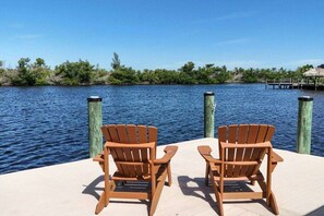 Two wooden Adirondack chairs on a dock with a view of a calm body of water and a forested shoreline. - CCVS - Villa Kara Tuscany (Cape Coral, FL)