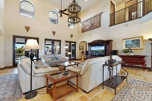 Un salon spacieux avec un plafond haut, un ventilateur de plafond, un grand téléviseur à écran plat et un balcon doté d’une balustrade.