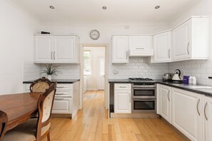 A kitchen with white cabinets, a wooden dining table, and a clock on the wall. - New! Charming 1BR Maisonette | Alexandra Park (Greater London)