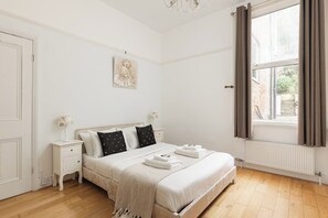 A bedroom with a bed, nightstands, a window with curtains, a radiator, and a picture on the wall. - New! Charming 1BR Maisonette | Alexandra Park (Greater London)