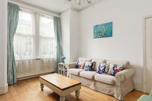 A living room with a wooden coffee table, a beige sofa with floral cushions, a blue painting on the wall, and a window with curtains. - New! Charming 1BR Maisonette | Alexandra Park (Greater London)