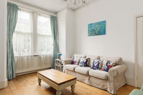 A living room with a wooden coffee table, a beige sofa with floral cushions, a blue painting on the wall, and a window with curtains. - New! Charming 1BR Maisonette | Alexandra Park (Greater London)
