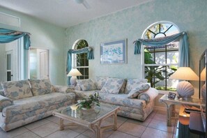 A living room with two sofas, a coffee table, and a lamp. There are two windows with curtains, a framed picture on the wall, and a door. - CCVS - Villa White Diamond (Cape Coral)