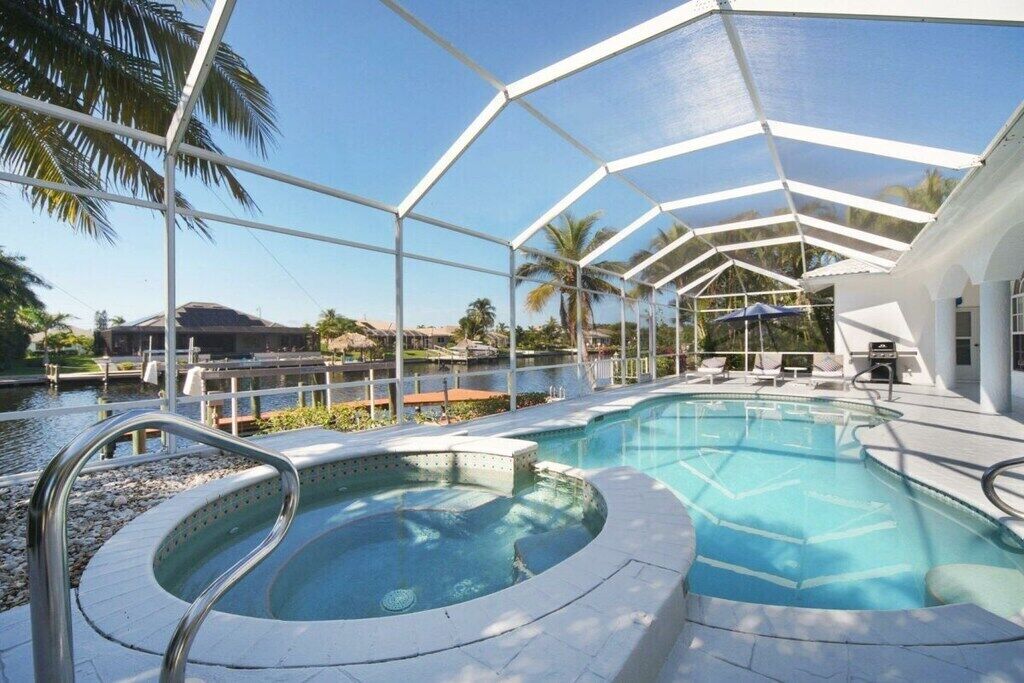 A swimming pool with a hot tub, surrounded by a covered area with palm trees and a view of a body of water.