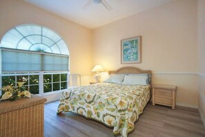 A bedroom with a large window, a bed with floral bedding, a bedside table, and a nightstand. - CCVS - Villa White Diamond (Cape Coral)