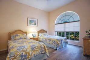 A bedroom with two beds, a window with a view of greenery, a painting on the wall, and a wicker side table with a lamp. - CCVS - Villa White Diamond (Cape Coral)