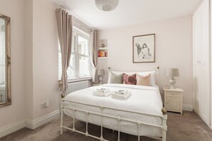 A bedroom with a white bed, a bedside table with a lamp, a radiator, a window with curtains, and a framed picture on the wall. - New! West Kensington 2BR Gem w/Garden & Patio (Greater London)