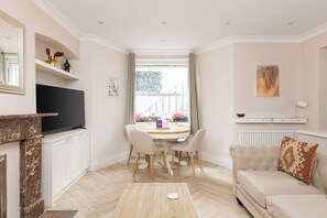 A living room with a fireplace, a television, a dining table with chairs, a sofa, and a window with curtains. - New! West Kensington 2BR Gem w/Garden & Patio (Greater London)