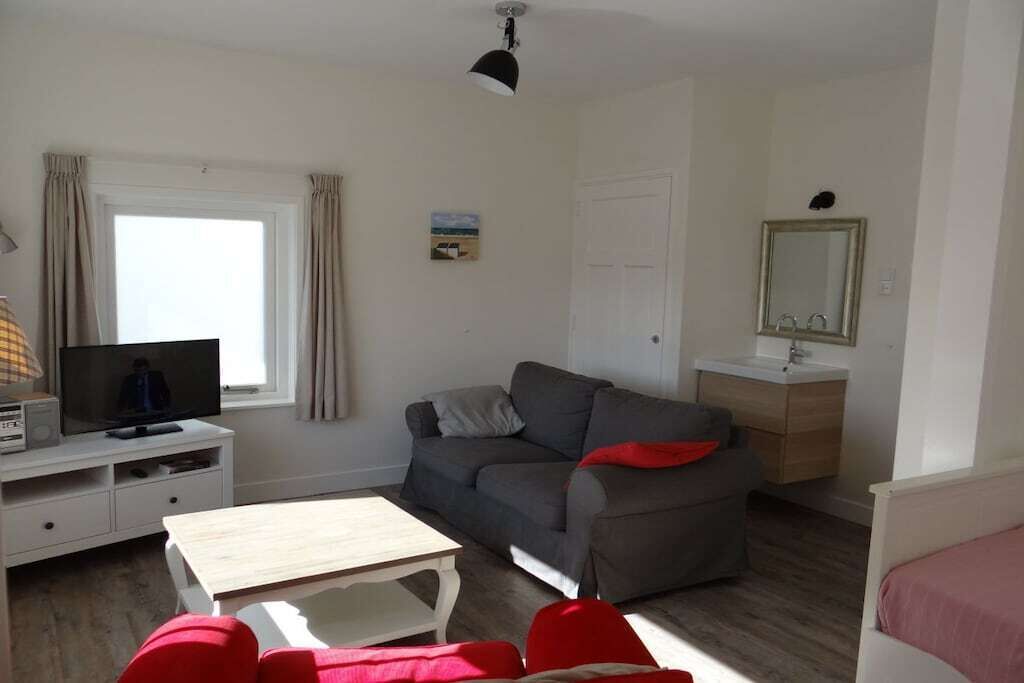 A compact living space with a sofa, a television, a small table, a bedside table, a mirror, and a window with curtains.