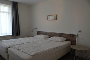 Ein Hotelzimmer mit zwei Betten, einem kleinen runden Tisch, einer Wandlampe und einem Fenster mit Vorhängen.