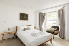 A bedroom with a bed, a bedside table, a chair, and a window with curtains. - New! Stylish 1BR Flat w/Patio in Vibrant Peckham (Greater London)
