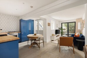 A modern kitchen with blue cabinets, a dining area with a round table, and a living space with a blue sofa and a TV. - New! Stylish 1BR Flat w/Patio in Vibrant Peckham (Greater London)