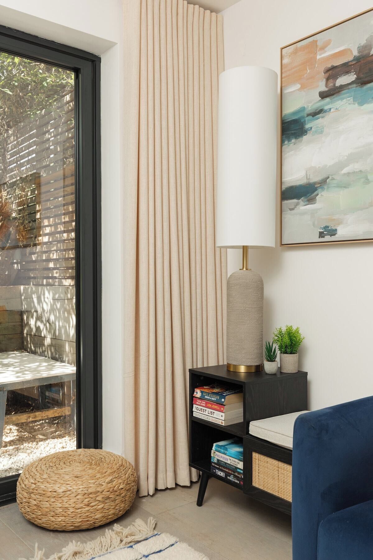 Un salon moderne avec une grande fenêtre, un canapé bleu, un pouf rond en fibres tressées, une bibliothèque noire remplie de livres, un grand lampadaire et un tableau accroché au mur.
