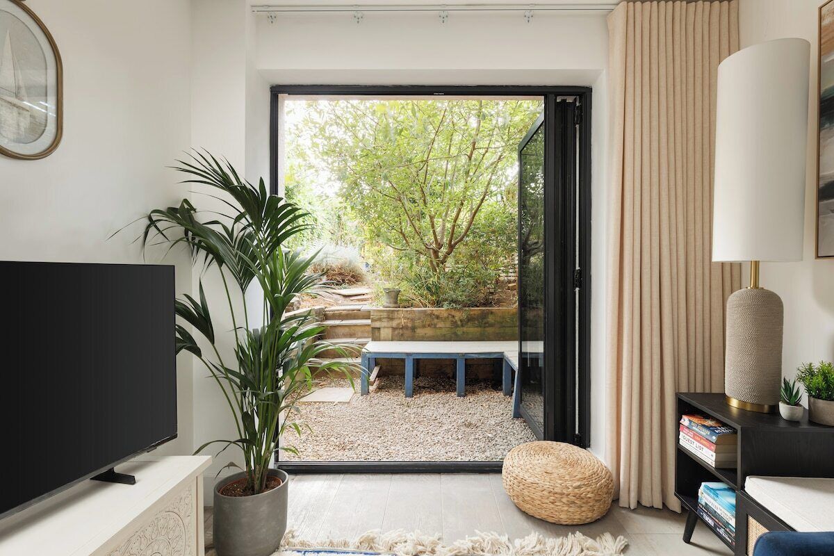 Una sala de estar moderna con televisor, planta en maceta, banco y vista a un jardín a través de un ventanal.