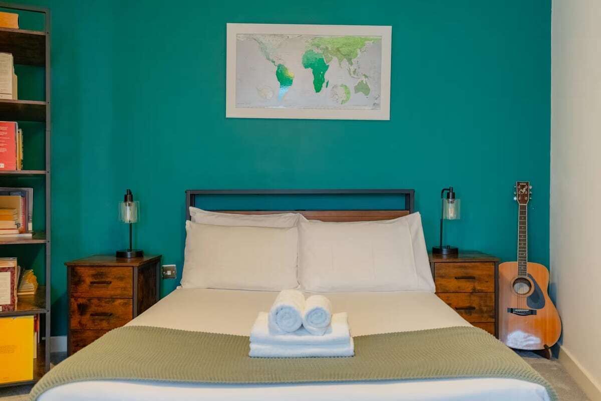 Un lit bien fait, avec des serviettes pliées, une carte du monde accrochée au mur, une guitare et une bibliothèque remplie de livres et surmontée d’une lampe.