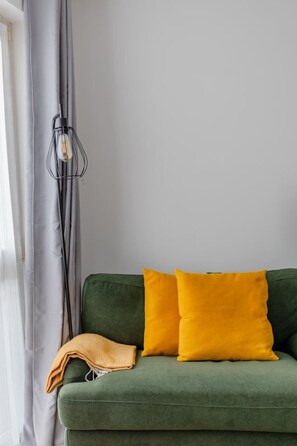 A green sofa with two yellow cushions and a single orange cushion, positioned against a white wall with a black light fixture. - New! Canal-side 1BR Flat in Clapton (Greater London)