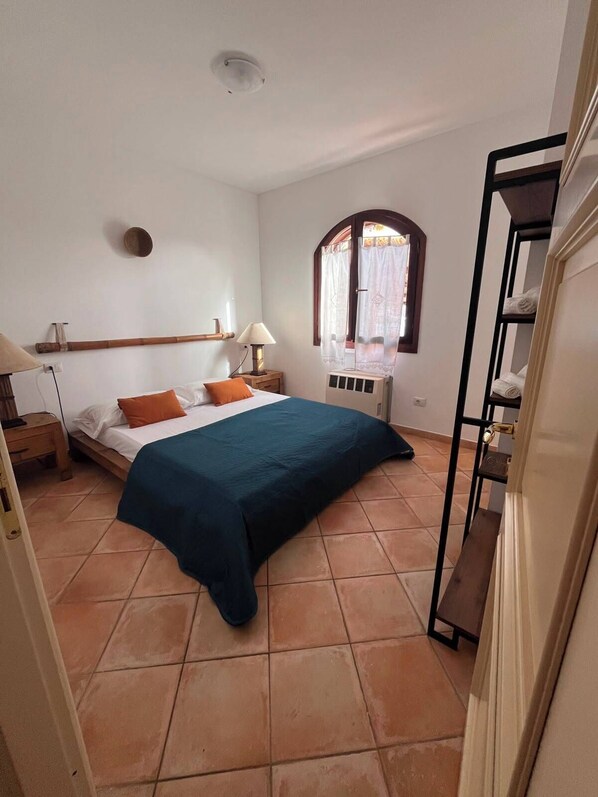 Una camera da letto con un letto, un comodino, una lampada, una finestra con tende e un termosifone.