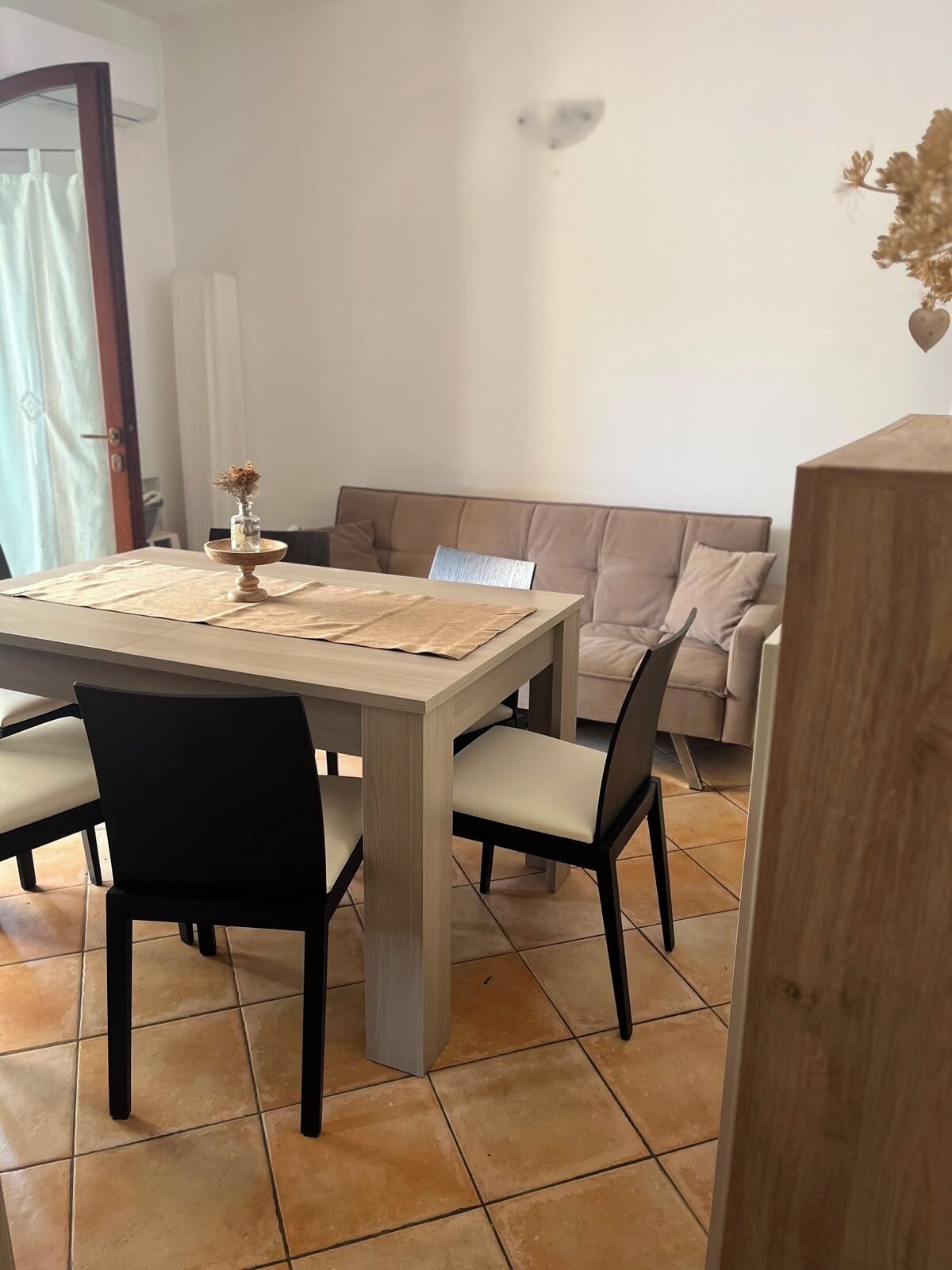 A dining area with a rectangular table, four chairs, a vase with dried flowers, a beige sofa, and a wall-mounted light fixture.