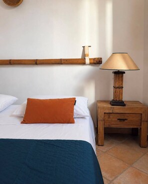 Un letto con lenzuola bianche e un cuscino arancione, un comodino in legno con una lampada e una mensola in bambù sopra il letto.