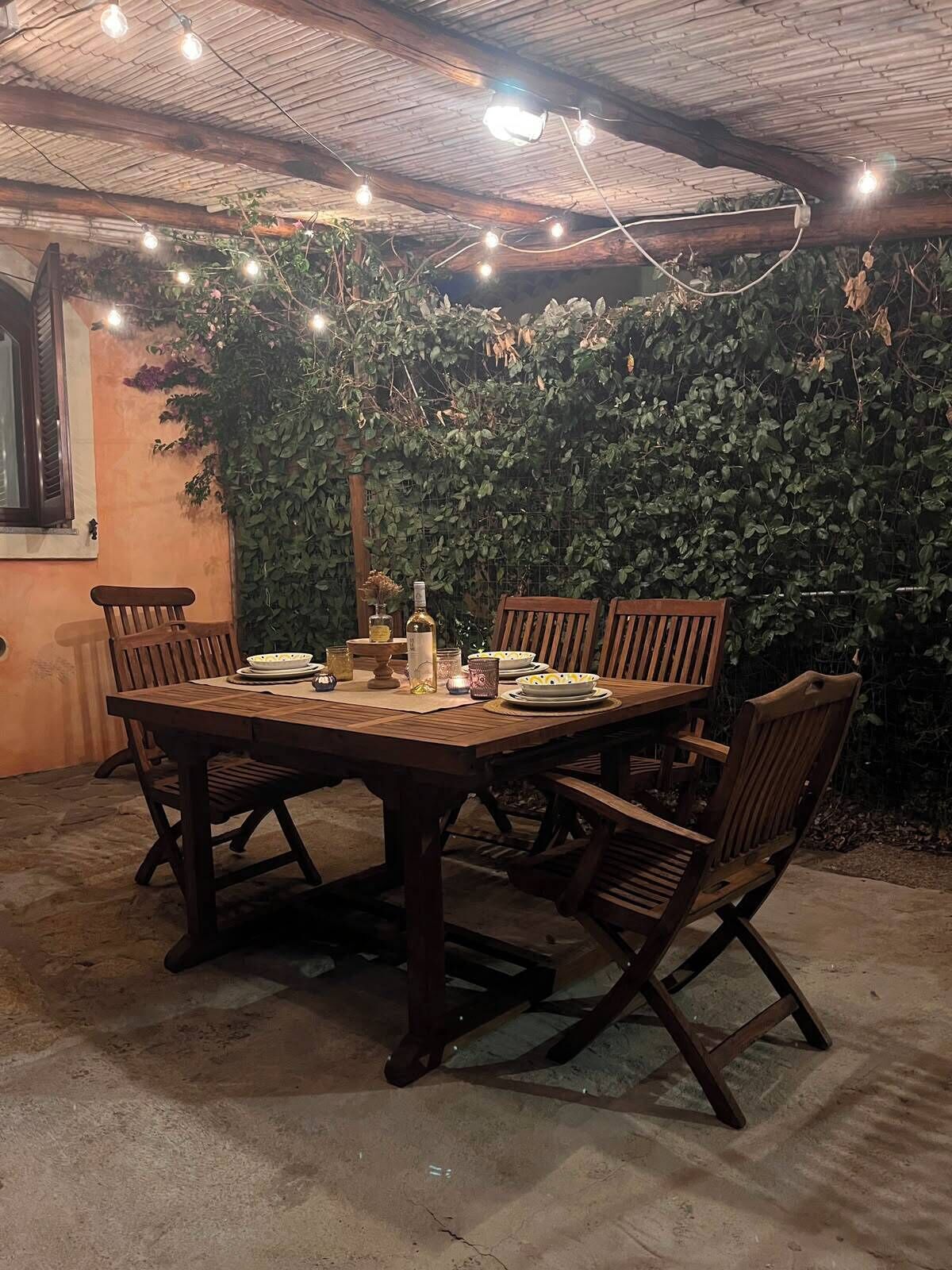 An outdoor dining area with a wooden table and chairs, surrounded by a lush green hedge.