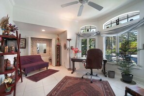 Un salon comprenant un canapé, un fauteuil, un bureau, un ventilateur de plafond et une fenêtre avec des stores.