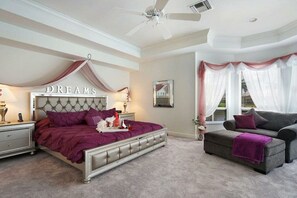 Ein Schlafzimmer mit einem gebügelten Kopfteil, einem Bett mit einer violetten Tagesdecke, einem grauen Sofa und einem Deckenventilator.