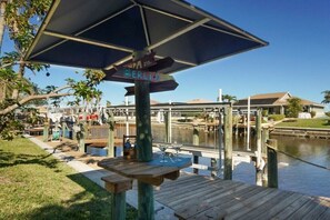 Eine Holzterrasse mit Tisch und Stühlen, ein Wegweiser mit Beschilderung und Blick auf einen Yachthafen mit Booten und einem Gebäude im Hintergrund.