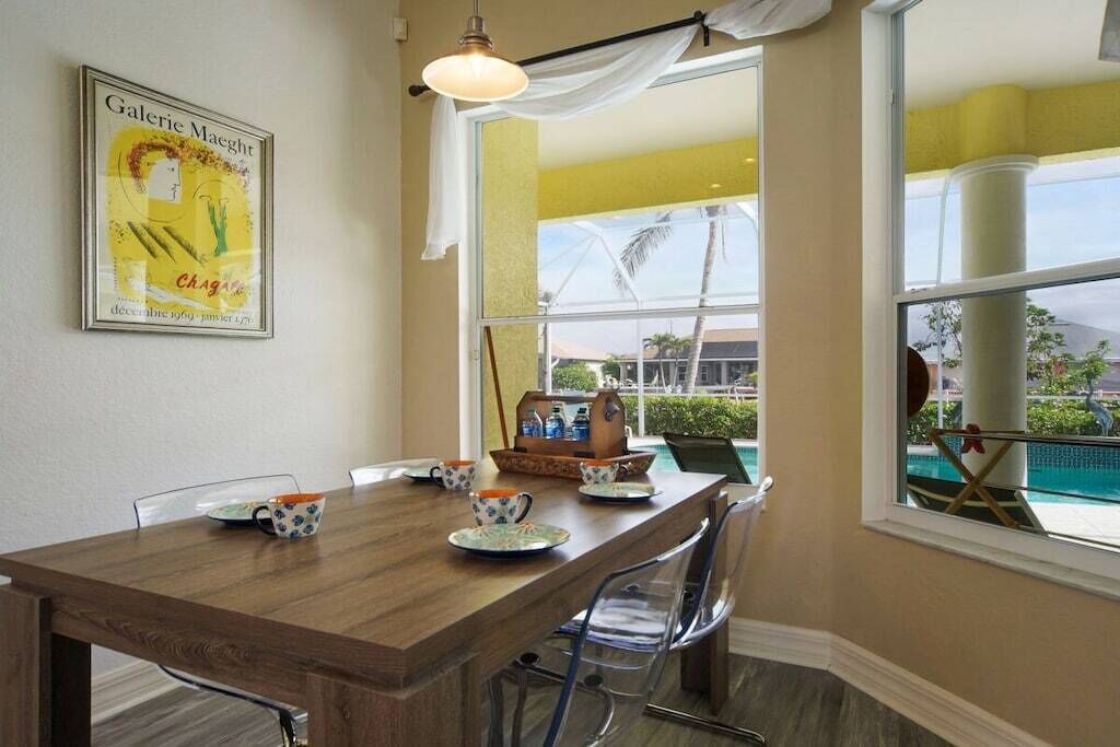 A dining area with a wooden table, chairs, a framed artwork, and a view of a pool through a large window.