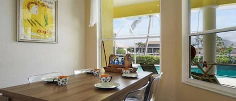 Un comedor con una mesa de madera, sillas, una obra de arte enmarco y vista a una piscina a través de un ventanal.