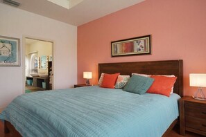 Ein Schlafzimmer mit einem großen Bett, zwei Nachttischen, einem Schminktisch und einem gerahmten Bild über dem Bett. Ein Badezimmer ist durch eine geöffnete Tür erkennbar.