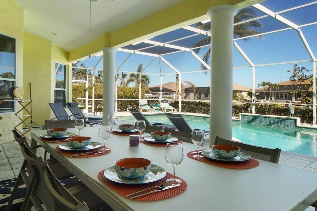 Mesa de comedor con platos, tazones y vasos, con vista a una piscina y sillones de descanso.
