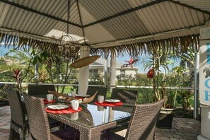 An outdoor dining area with a glass table, wicker chairs, and a thatched roof. - CCVS - Villa Sunny Breeze (Cape Coral)