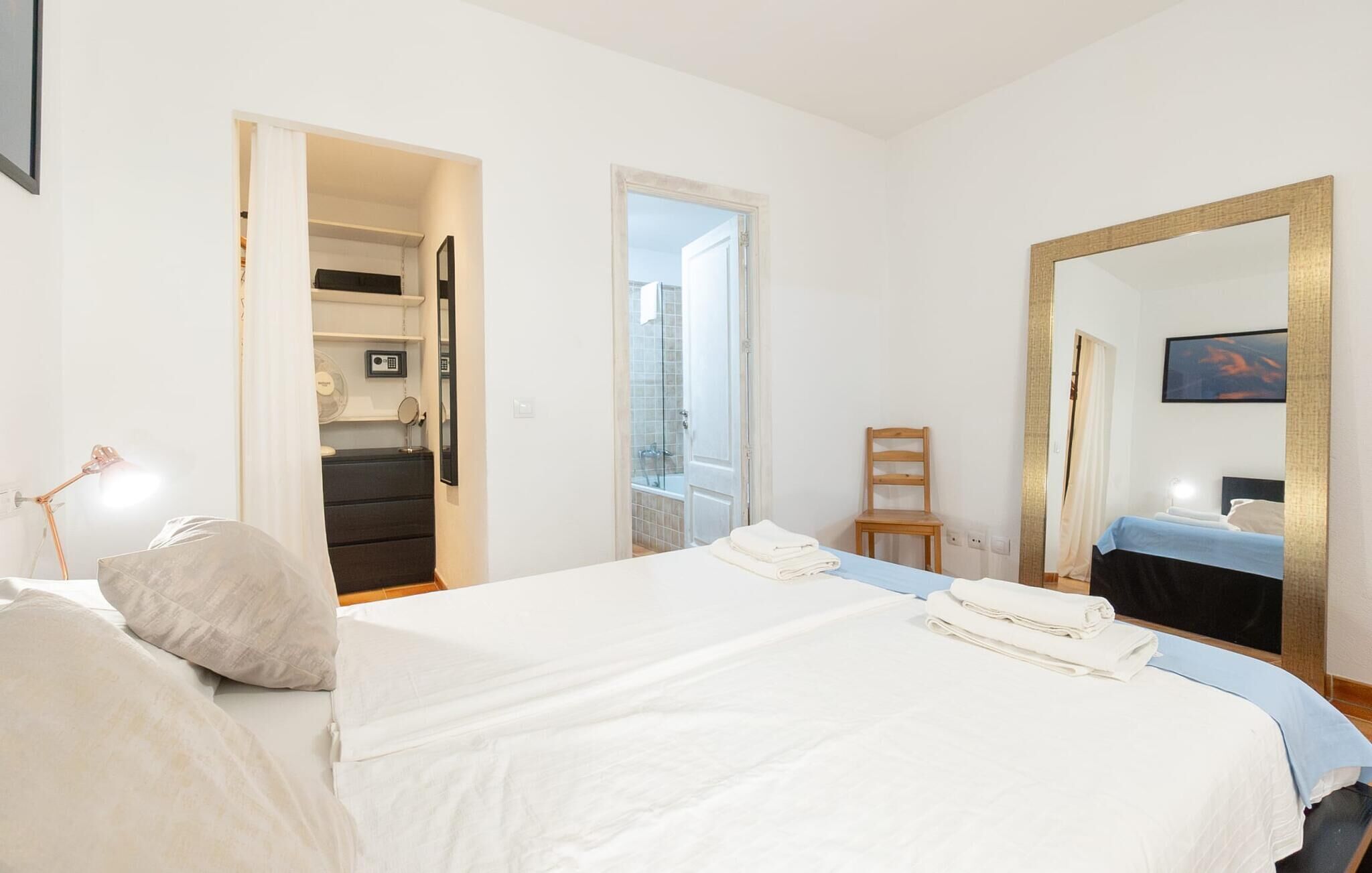 A hotel room with a bed, a chair, a desk with a lamp, a mirror, and a bathroom visible through an open door.