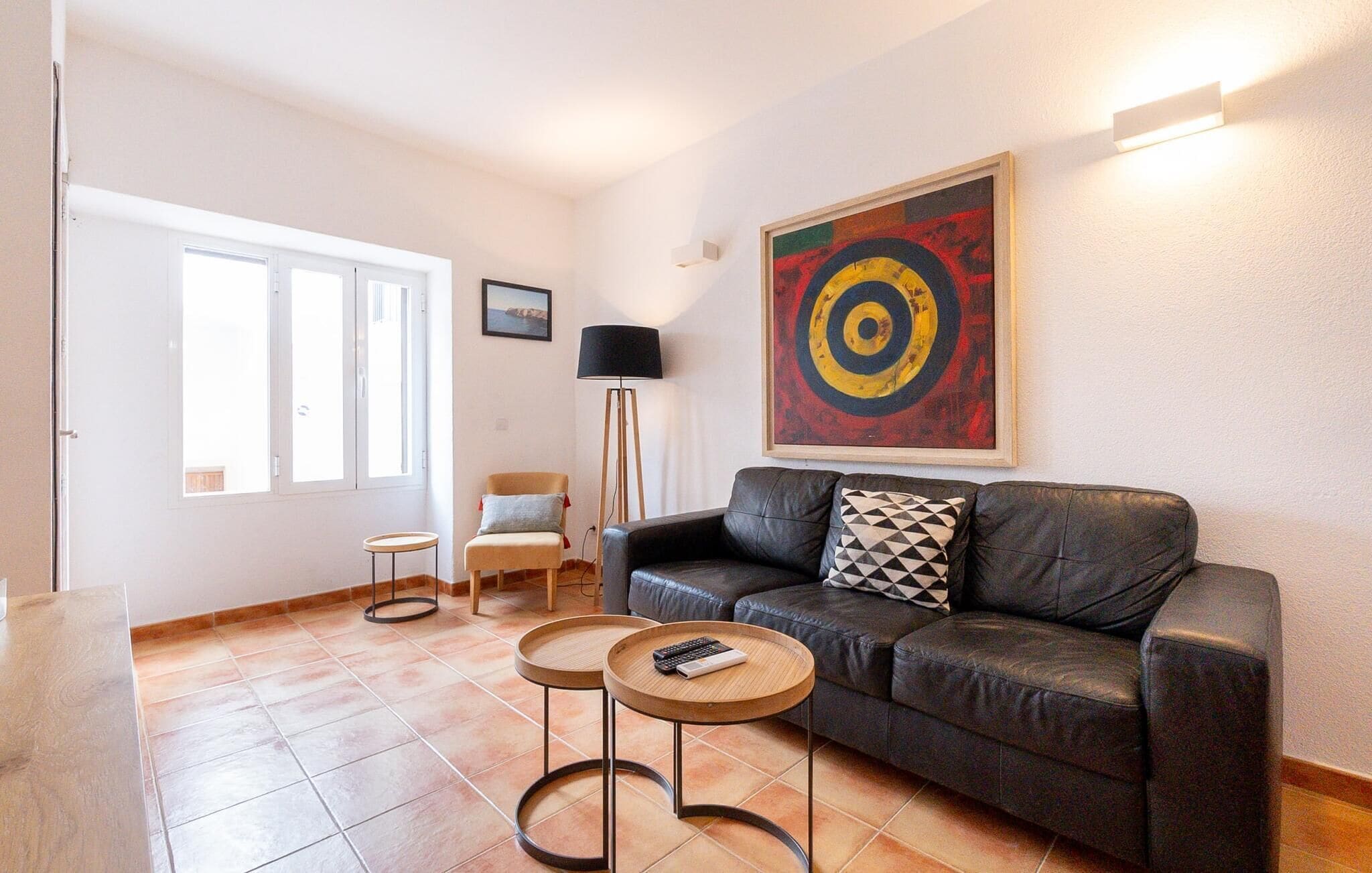 A living room with a leather sofa, two small round tables, a floor lamp, and a large abstract painting on the wall.