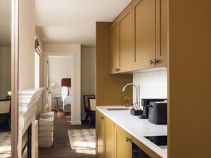 Cocina con encimera blanca, un lavamanos y armarios. Se ve una zona de comedor con una mesa y sillas a través de una puerta abierta.