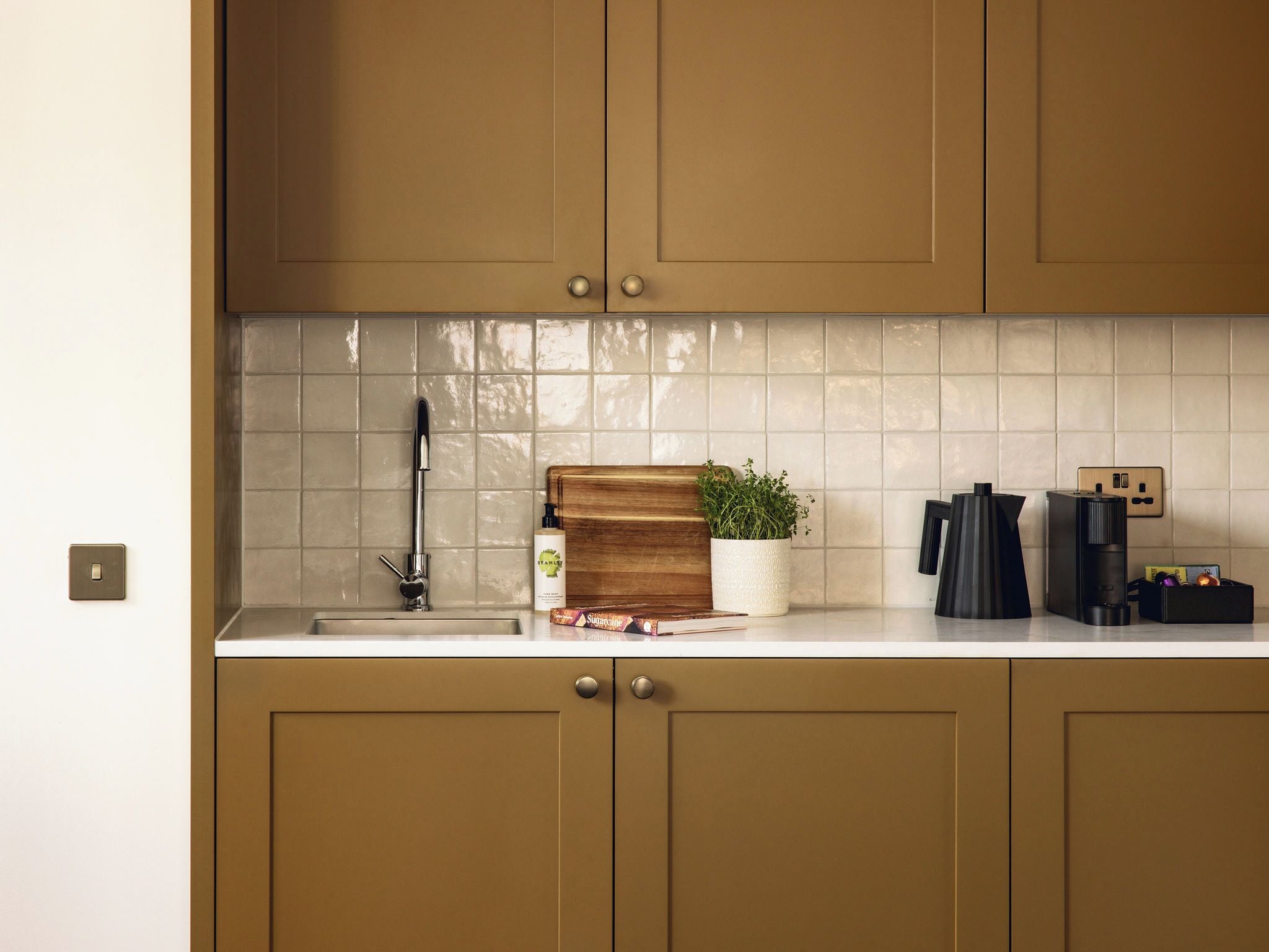 A kitchen with a white countertop, a wooden cutting board, a potted plant, a coffee maker, and a book.