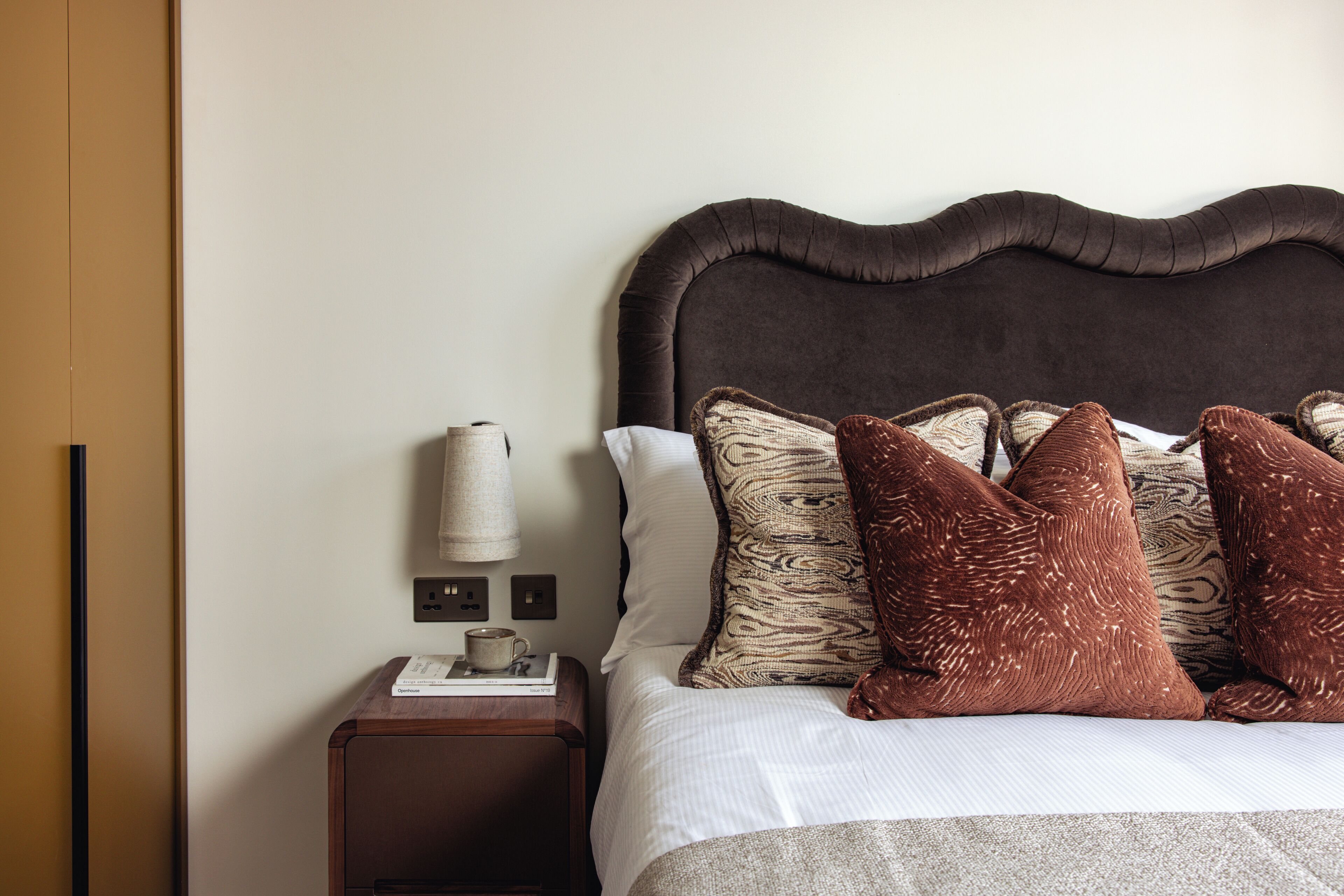 A neatly made bed with a dark headboard, a bedside table with a lamp, and a power outlet.