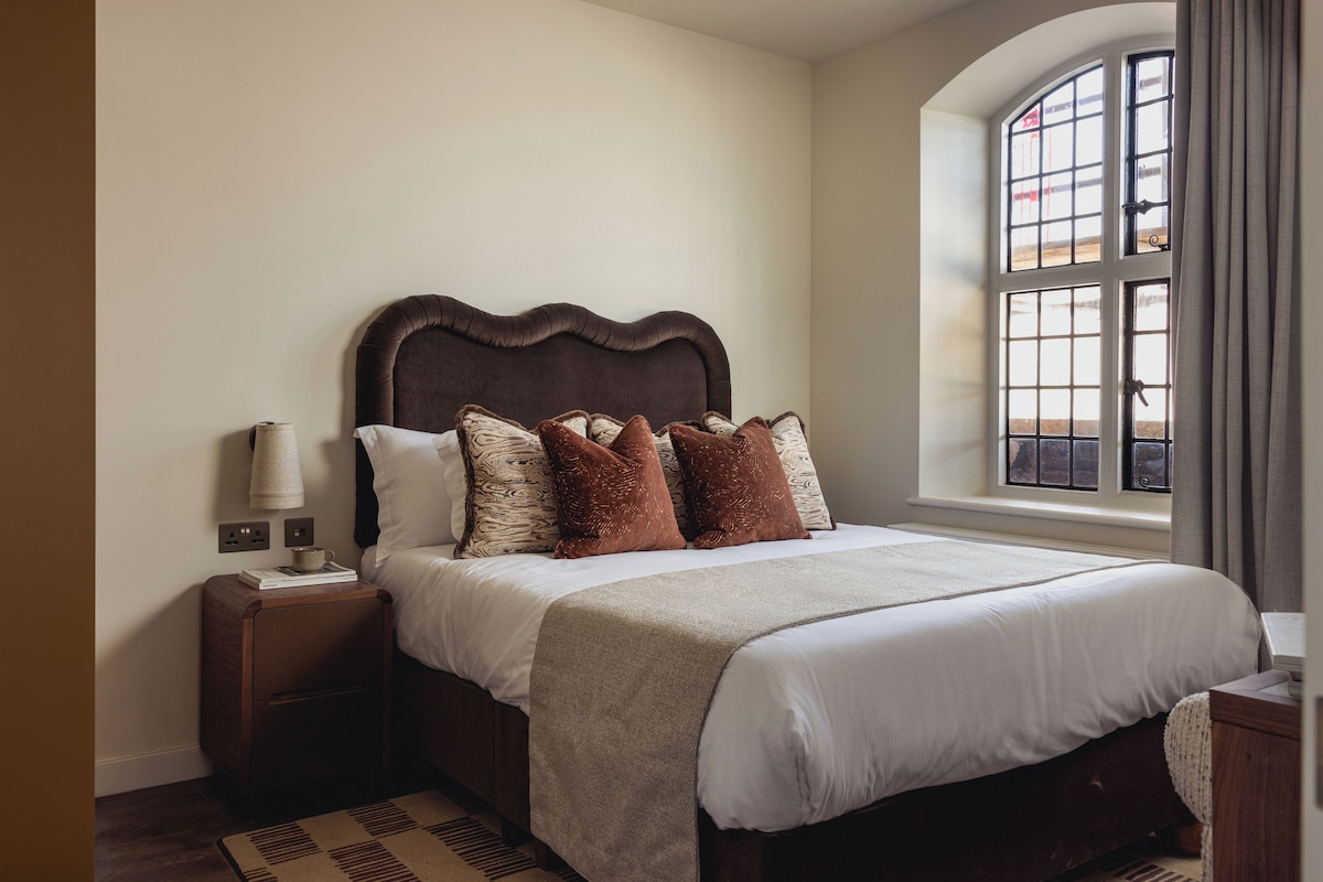 Un dormitorio con una cama grande, una mesita de noche, una lámpara y una ventana con cortinas.