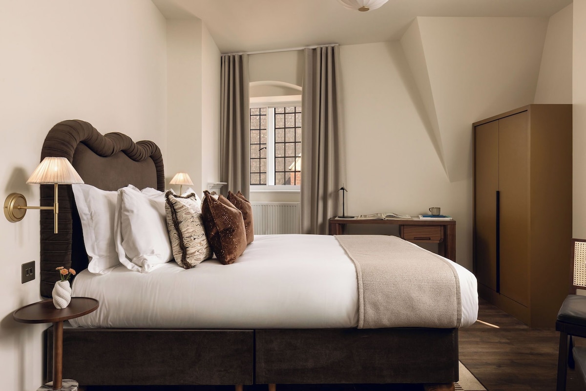 Un dormitorio moderno con una cama grande, lámparas de noche, una ventana con cortinas, un armario de madera y una mesita con un libro y una taza.