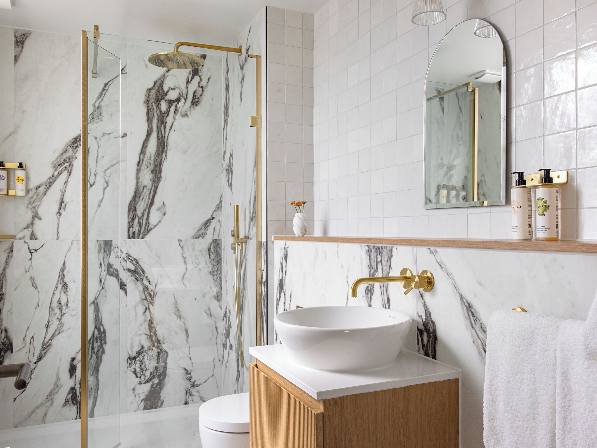 Un baño con pared de mármol, un lavamanos blanco, un grifo dorado, una ducha con cabezal dorado y un espejo sobre el lavamanos.