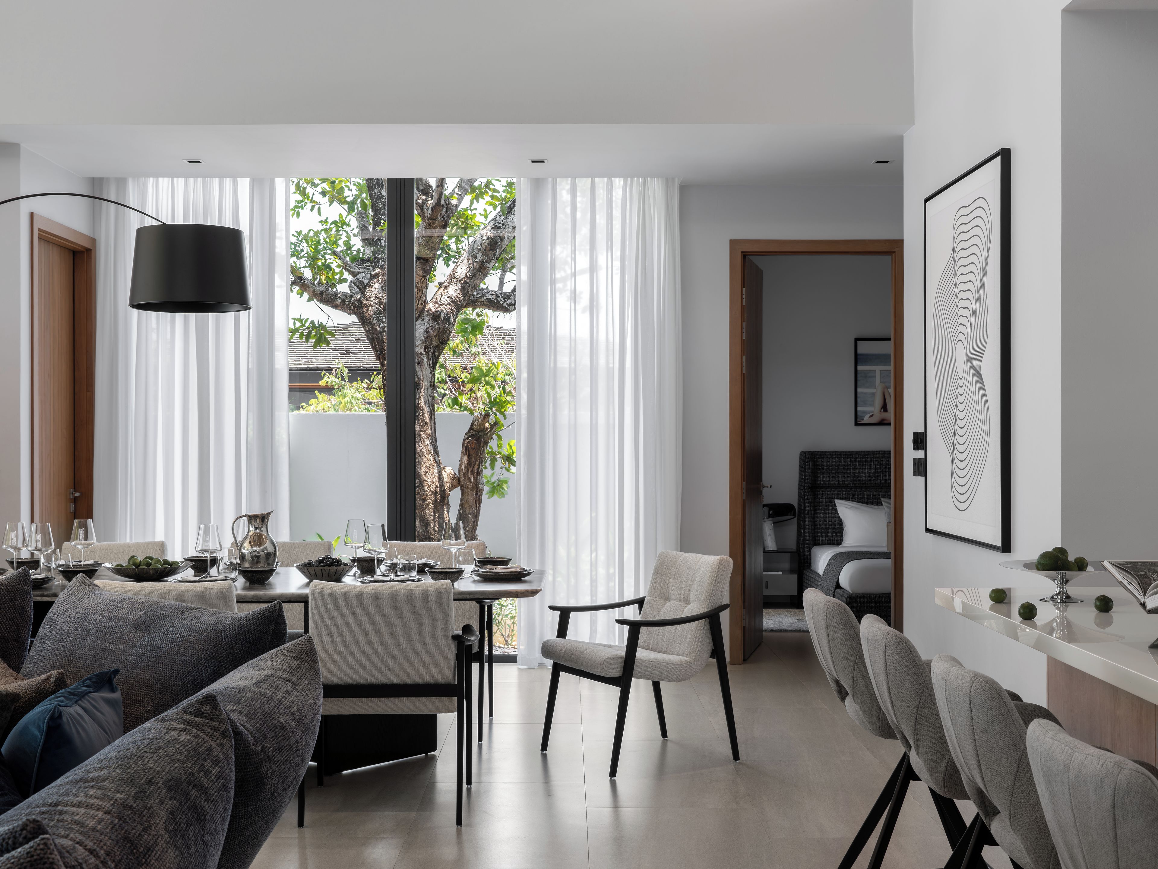 A modern living room with a dining area, a large window with curtains, a tree outside, and a framed artwork on the wall.