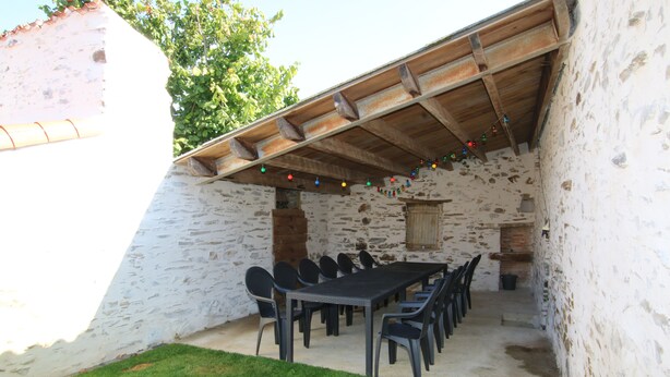 Uma área de refeições ao ar livre rústica, com parede de pedra, teto de madeira e uma mesa comprida com cadeiras.