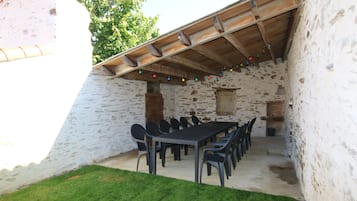 A rustic outdoor dining area with a stone wall, wooden ceiling, and a long table with chairs.
