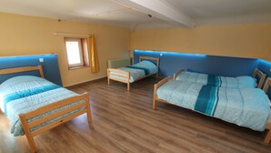 Quarto com três camas de madeira, cada uma com colcha estampada em azul e branco. O quarto possui piso de madeira e uma janela com cortina amarela.