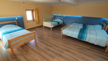 A room with three wooden beds, each with a blue and white patterned bedspread. The room has wooden flooring and a window with a yellow curtain.